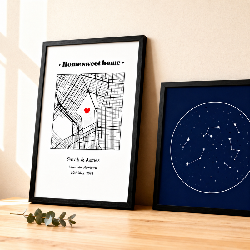 Framed map with a red heart and 'Home sweet home' text on a white background, next to a framed star constellation chart on a wooden surface.