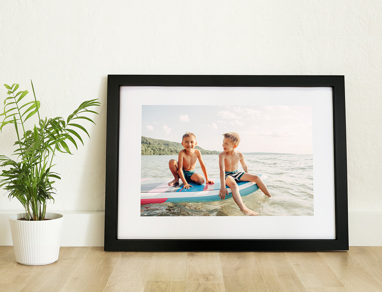 A framed print with a photo inside featuring two children sitting on a surfboard, displayed on a wall with a white mount and a black frame, placed next to a potted plant.