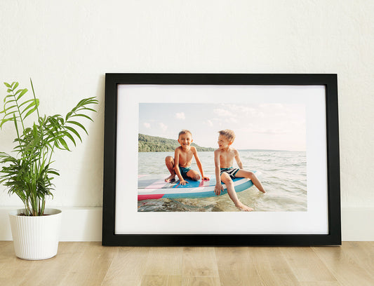 A framed print with a photo inside featuring two children sitting on a surfboard, displayed on a wall with a white mount and a black frame, placed next to a potted plant.
